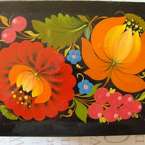 Op de afbeelding: Een rechthoekige zwarte houten doos met een levendig bloemenontwerp. De bloemen zijn geschilderd in tinten rood, oranje en geel, met groene bladeren en blauwe accenten. De doos heeft een glanzende afwerking.