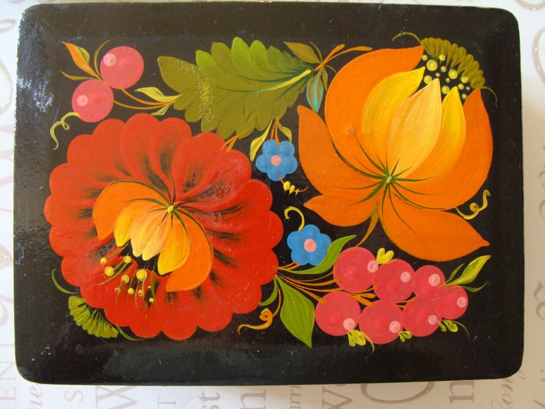 Op de afbeelding: Een rechthoekige zwarte houten doos met een levendig bloemenontwerp. De bloemen zijn geschilderd in tinten rood, oranje en geel, met groene bladeren en blauwe accenten. De doos heeft een glanzende afwerking.