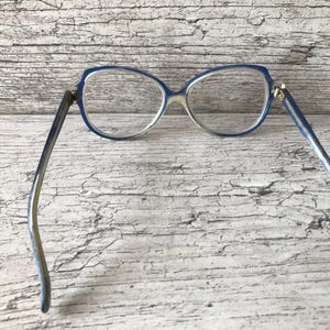 May include: Vintage spectacles with a blue frame and clear lenses. The glasses have a cat-eye shape and a light blue and silver-toned arm. The frame is made of plastic and the lenses are clear. The glasses are set against a weathered wood background.