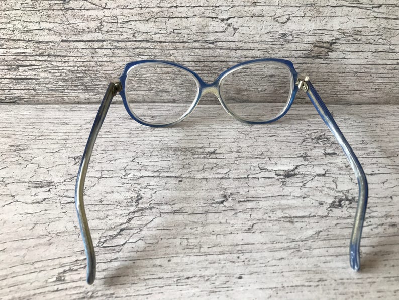 May include: Vintage spectacles with a blue frame and clear lenses. The glasses have a cat-eye shape and a light blue and silver-toned arm. The frame is made of plastic and the lenses are clear. The glasses are set against a weathered wood background.