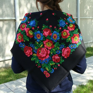 May include: A black square scarf featuring a vibrant floral pattern. The design includes large red, pink, and yellow flowers with blue accents and green leaves. The scarf is draped over the shoulders.