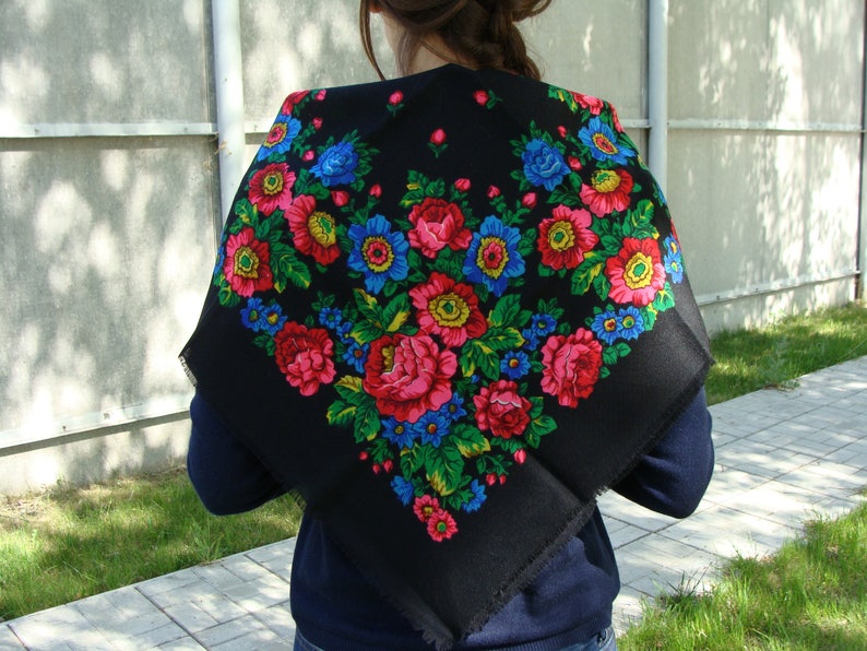 May include: A black square scarf featuring a vibrant floral pattern. The design includes large red, pink, and yellow flowers with blue accents and green leaves. The scarf is draped over the shoulders.