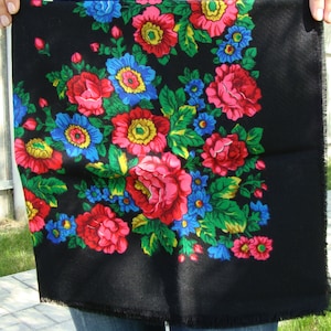 May include: Black square scarf with a vibrant floral pattern. The design features large red, pink, and blue flowers with green leaves against a black background. The scarf is held up, showing its fringed edges.
