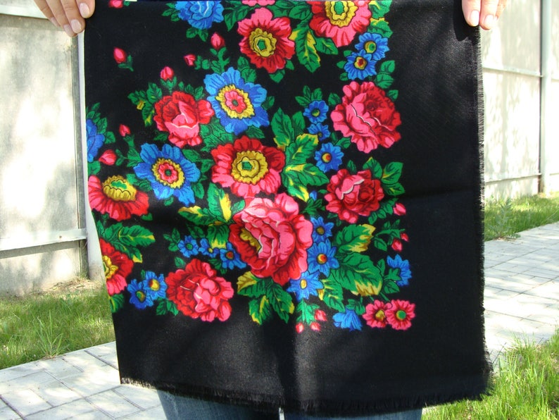 May include: Black square scarf with a vibrant floral pattern. The design features large red, pink, and blue flowers with green leaves against a black background. The scarf is held up, showing its fringed edges.