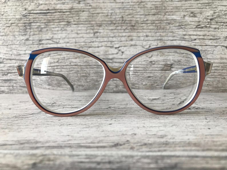 May include: Vintage spectacles with a rounded, oversized frame. The frame is mainly a muted rose colour, with a thin blue stripe along the top edge. The temples are silver-toned, and the lenses are clear.