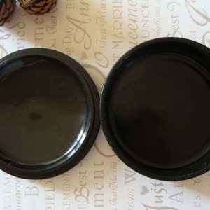 May include: Two open, round, black containers with a glossy finish. The containers are placed side by side, revealing their interior. The background features text and pinecones.