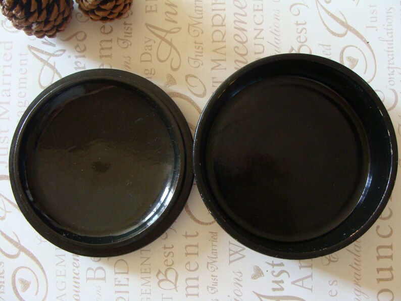 May include: Two open, round, black containers with a glossy finish. The containers are placed side by side, revealing their interior. The background features text and pinecones.