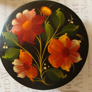 May include: A round, black decorative box with a hand-painted floral design. The flowers are in shades of red, orange, and yellow, with green leaves and white accents. The box has a glossy finish and a traditional folk art style.