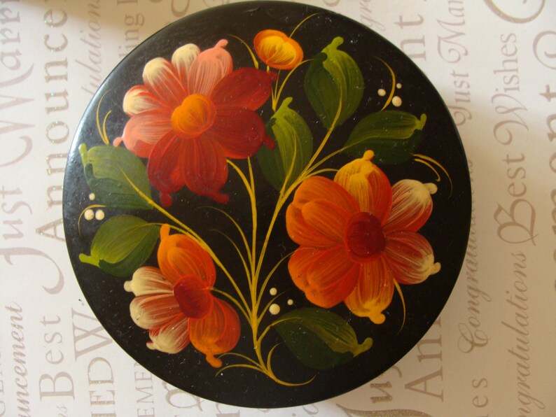 May include: A round, black decorative box with a hand-painted floral design. The flowers are in shades of red, orange, and yellow, with green leaves and white accents. The box has a glossy finish and a traditional folk art style.