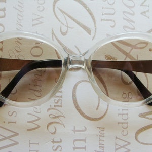 May include: Clear, oversized sunglasses with brown-tinted lenses. The frames are transparent, with black arms and a gold-colored bridge. These vintage-style glasses offer a retro aesthetic.