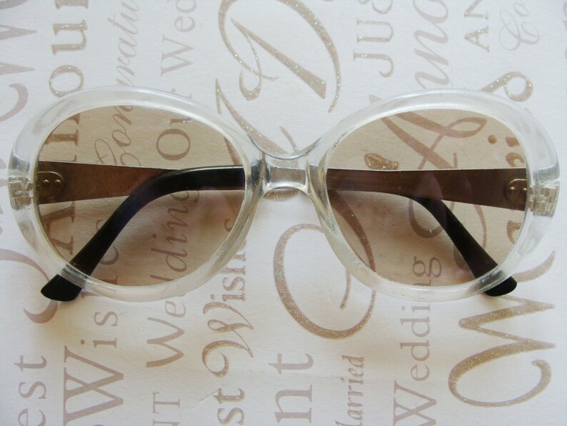 May include: Clear, oversized sunglasses with brown-tinted lenses. The frames are transparent, with black arms and a gold-colored bridge. These vintage-style glasses offer a retro aesthetic.