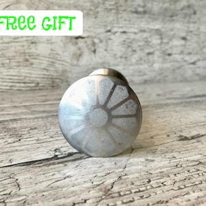 Peut inclure: Un bouton de meuble argenté avec un motif de soleil. Le bouton est rond et a une finition métallique. Le texte "+ FREE GIFT" est en vert en haut à gauche.
