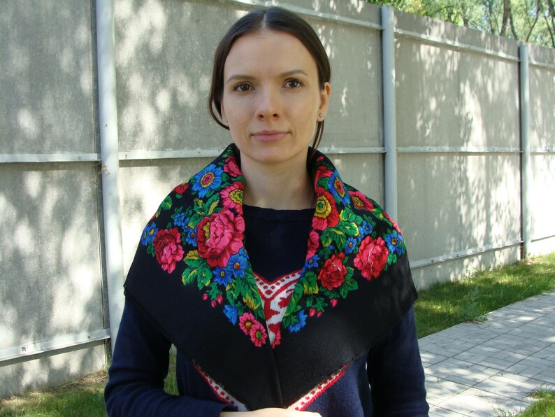 May include: A black shawl with a vibrant floral pattern featuring pink, red, blue, and yellow flowers. The shawl is worn around the neck and shoulders, creating a colorful contrast against the dark background.