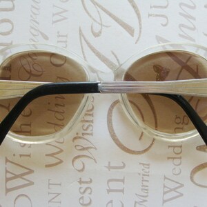 May include: Clear oval-shaped sunglasses with brown lenses and silver-toned metal accents. The frames have a retro design with black temples. A stylish accessory for eye protection.