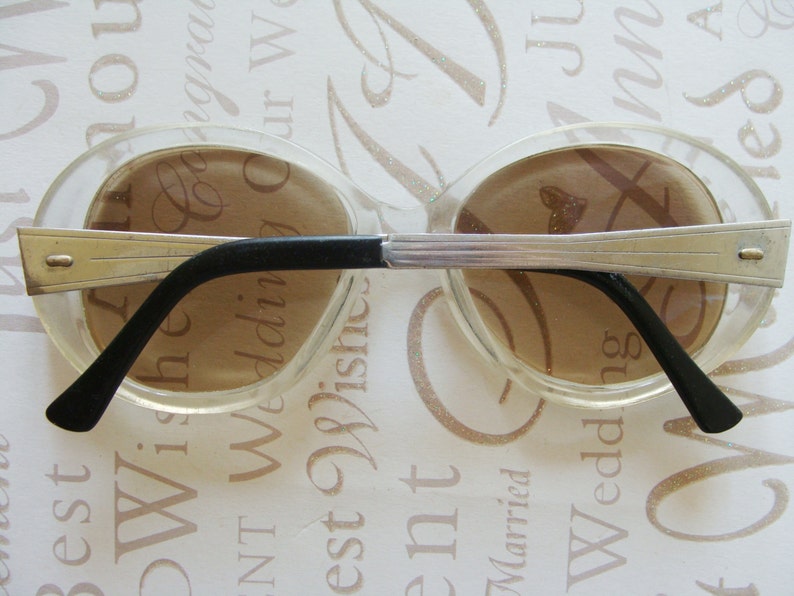 May include: Clear oval-shaped sunglasses with brown lenses and silver-toned metal accents. The frames have a retro design with black temples. A stylish accessory for eye protection.
