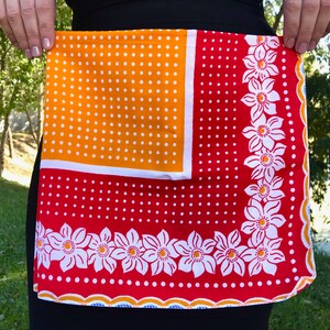 May include: A colourful apron with a retro design. The apron features a vibrant orange polka-dot top section, a red lower section with white polka dots, and a floral border. The apron is held up by a person.