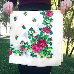 May include: Cream-coloured square scarf with a floral design featuring large red and pink roses, green leaves, and small red buds. The scarf is held up, showcasing the detailed pattern and colour contrast.