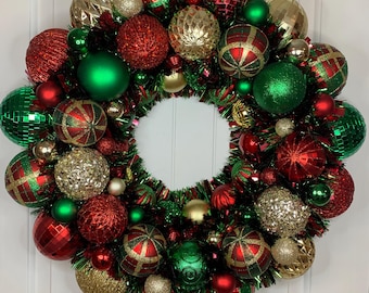 May include: A festive Christmas wreath composed of red, green, and gold ornaments. The wreath features a variety of ball shapes, including glittery and patterned designs, arranged around a central opening. Green tinsel adds a touch of sparkle.