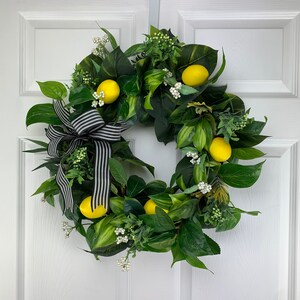 Puede incluir: Una corona verde con limones y flores blancas, decorada con una cinta a rayas blancas y negras.