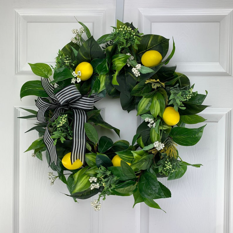 Puede incluir: Una corona verde con limones y flores blancas, decorada con una cinta a rayas blancas y negras.