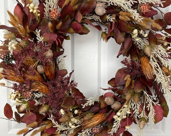 May include: A circular wreath featuring a variety of dried flowers and foliage. The wreath has deep red, brown, and cream-colored elements, including leaves, seed pods, and feathery grasses. The wreath is displayed against a white door.