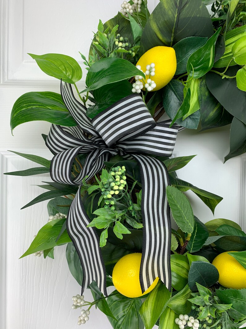 Puede incluir: Una corona de hojas verdes y limones amarillos est&aacute; decorada con un lazo de cinta a rayas blancas y negras.