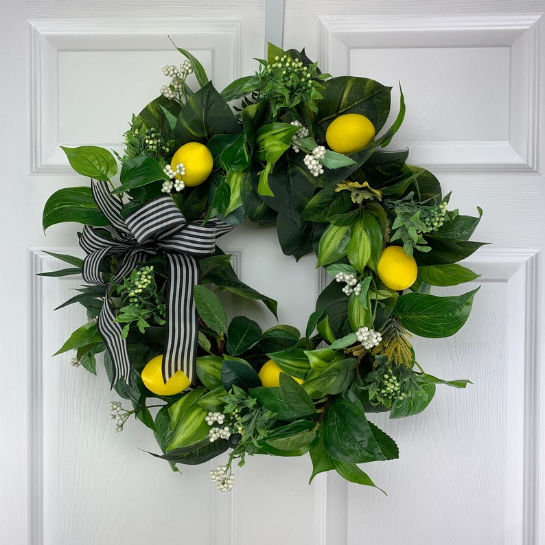 Puede incluir: Una corona verde y blanca con un lazo a rayas blanco y negro. La corona est&aacute; decorada con limones y flores blancas.