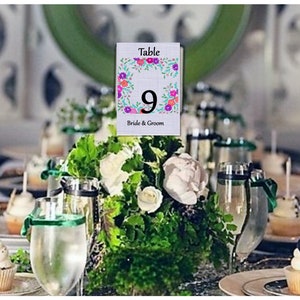 Könnte beinhalten: Eine weiße Tischnummerkarte mit einem floralen Rand und der Nummer 9 in Rosa und Lila. Die Karte trägt die Aufschrift "Tisch" und "Braut & Bräutigam".