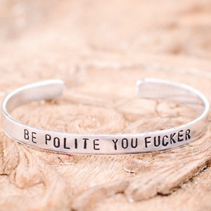 May include: A silver cuff bracelet with the stamped text "BE POLITE YOU FUCKER". The bracelet is open-ended and sits on a wooden surface. The text is in a bold, sans-serif font.