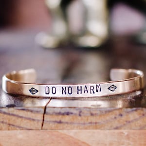May include: A gold-toned metal bracelet with the inscription "DO NO HARM" and arrow symbols on either side.
