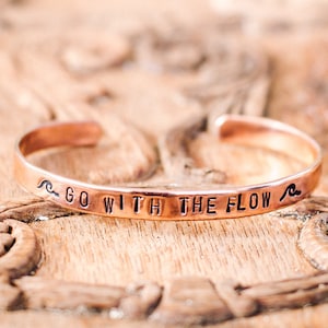 May include: A copper bracelet with the words "Go with the flow" engraved on it. The bracelet is decorated with two small wave symbols on either side of the text.