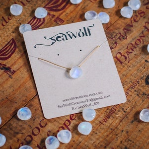 May include: A delicate gold necklace with a moonstone pendant on a wooden surface with the text "Seawolf" and contact information for the seller.