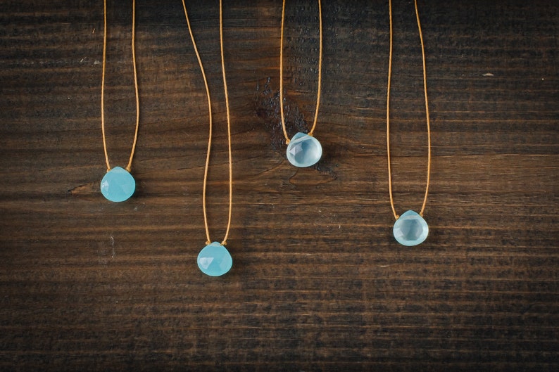 May include: Four delicate necklaces with light blue gemstone pendants hang on a brown wooden surface. Each necklace features a simple, thin, gold cord.