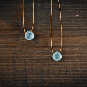 May include: Two delicate necklaces with light blue teardrop-shaped gemstones hanging from thin brown cords. The necklaces are displayed against a dark brown wooden background.