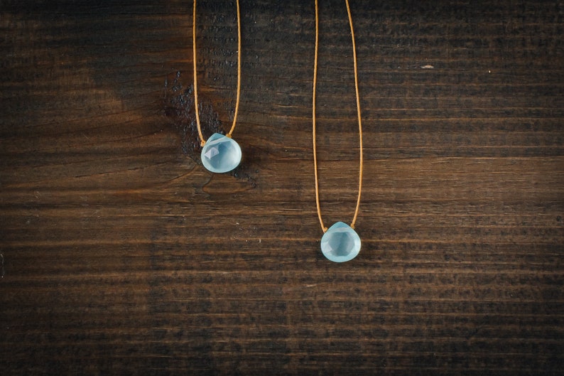 May include: Two delicate necklaces with light blue teardrop-shaped gemstones hanging from thin brown cords. The necklaces are displayed against a dark brown wooden background.