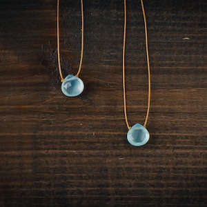 May include: Two delicate necklaces with light blue teardrop-shaped stones hanging from thin gold cords. The necklaces are displayed against a dark brown wooden background.