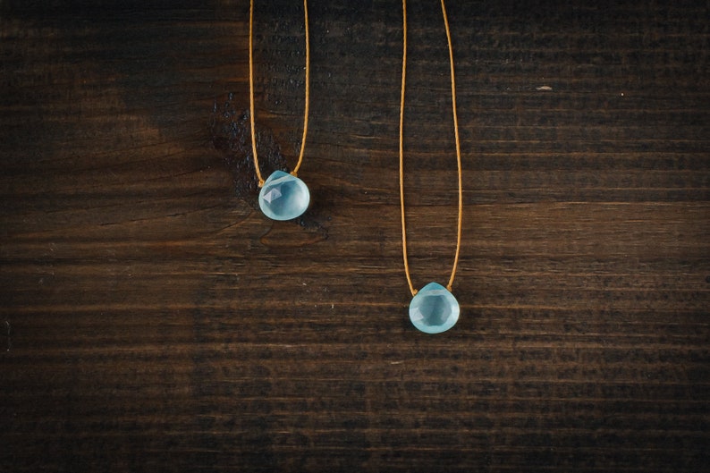 May include: Two delicate necklaces with light blue teardrop-shaped stones hanging from thin gold cords. The necklaces are displayed against a dark brown wooden background.