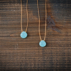 May include: Two delicate necklaces with light blue teardrop-shaped stones hanging from thin brown cords. The necklaces are displayed against a dark brown wooden background.