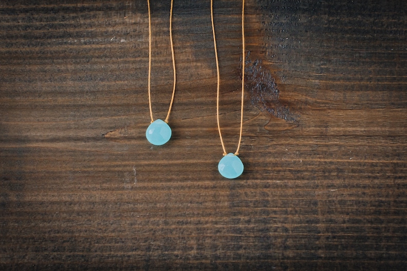 May include: Two delicate necklaces with light blue teardrop-shaped stones hanging from thin brown cords. The necklaces are displayed against a dark brown wooden background.