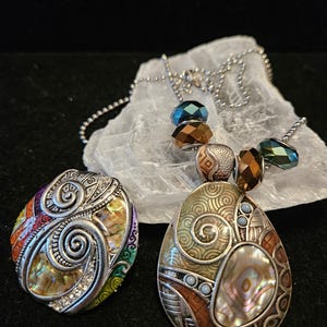 May include: Two decorative necklaces with pendants. One pendant is oval-shaped with a mother-of-pearl center and intricate metalwork. The other is round with a rainbow-colored design. Both have silver-tone chains and colorful bead accents.