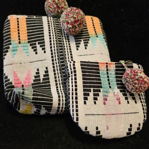 May include: Two fabric pouches with a black and white geometric pattern, accented with orange, teal, and pink. Each pouch is topped with a colorful pom-pom. The pouches are likely used for storing small items.