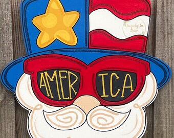 May include: A wooden door hanger shaped like a cartoon face wearing a red, white, and blue top hat with a star on it and sunglasses that say "AMERICA".