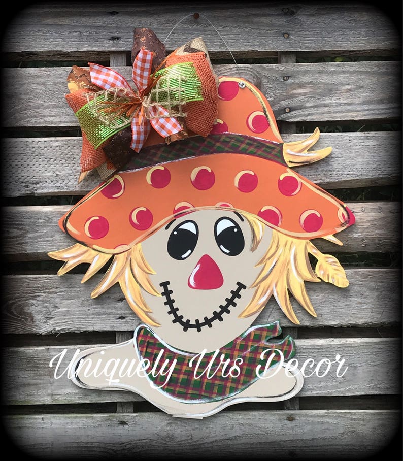 Peut inclure: Une d&eacute;coration de porte en bois peinte en forme de t&ecirc;te d'&eacute;pouvantail avec un grand chapeau orange &agrave; pois blancs, un nez rouge et un sourire cousu. L'&eacute;pouvantail porte une &eacute;charpe &agrave; carreaux marrons et verts et un grand n&oelig;ud avec un ruban &agrave; carreaux orange, vert et blanc.