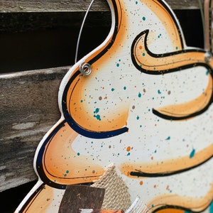 May include: A wooden cutout of a whipped cream topping with orange and brown accents. The cutout is decorated with white, brown, and teal paint splatters. The cutout is attached to a burlap bow with a pumpkin print.