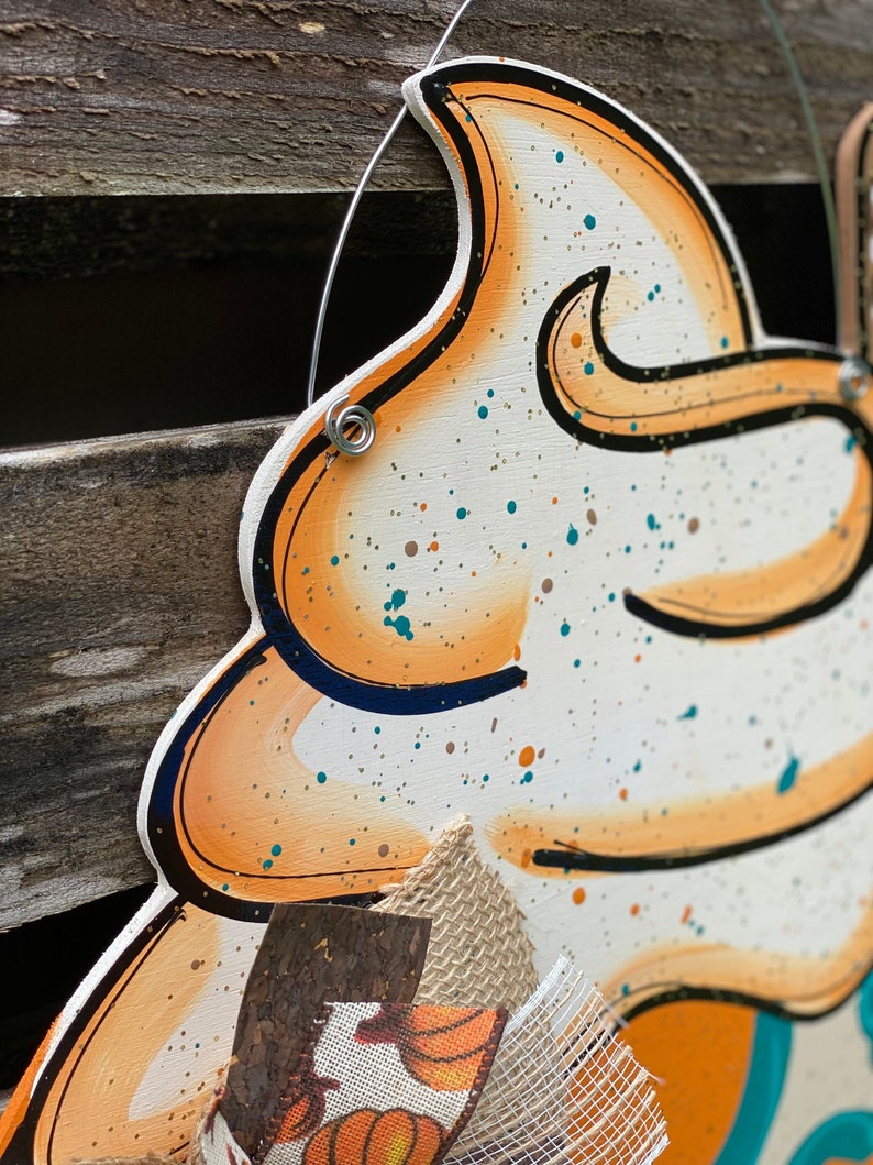 May include: A wooden cutout of a whipped cream topping with orange and brown accents. The cutout is decorated with white, brown, and teal paint splatters. The cutout is attached to a burlap bow with a pumpkin print.