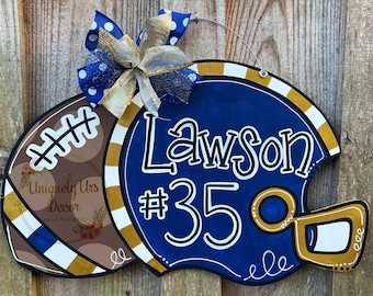 May include: A blue and gold football helmet door hanger with the name "Lawson" and the number "#35" painted on it. The helmet is decorated with white and gold stripes. A brown football with white and gold stripes is attached to the helmet.
