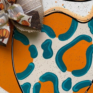 May include: A wooden pumpkin door hanger with a teal and orange abstract pattern. A burlap bow with a brown and white ribbon and small pumpkins is attached to the top.
