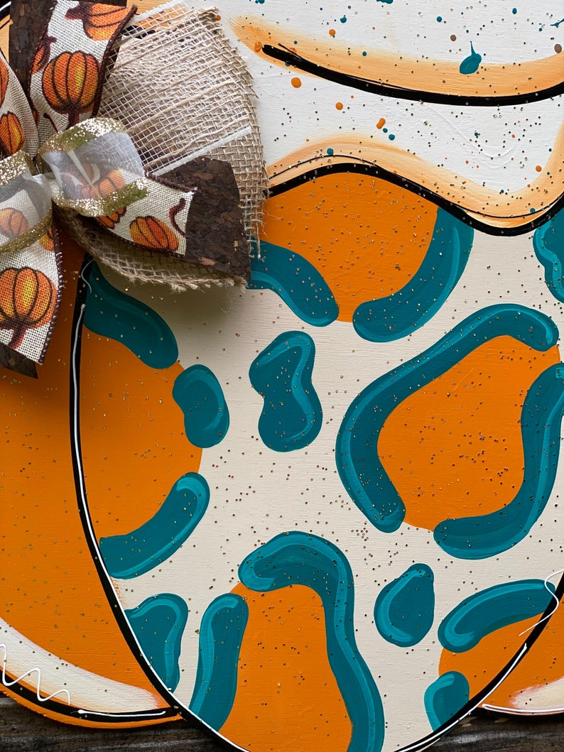 May include: A wooden pumpkin door hanger with a teal and orange abstract pattern. A burlap bow with a brown and white ribbon and small pumpkins is attached to the top.