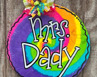 Può includere: Un appendiporta rotondo in legno con un motivo tie-dye in tonalità di viola, blu, verde, giallo e arancione. Le parole "Mrs. Dady" sono scritte in bianco sull'appendiporta. Un fiocco colorato con pois rosa, gialli, verdi e bianchi e motivi a quadri è attaccato alla parte superiore dell'appendiporta.
