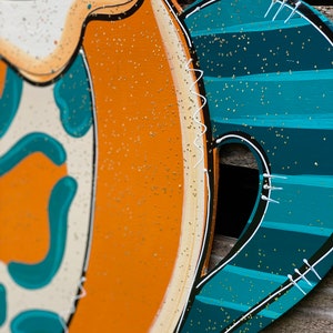 May include: A wooden door hanger shaped like a pumpkin with a teal and white striped design. The pumpkin is decorated with gold glitter and white stitching.
